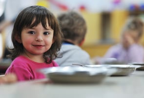 A young white girl with brunette hair wearing a pink shirt smiling at camera from the left side with a silver plate in front of her. The back of a blond boy's head is defocused in the background