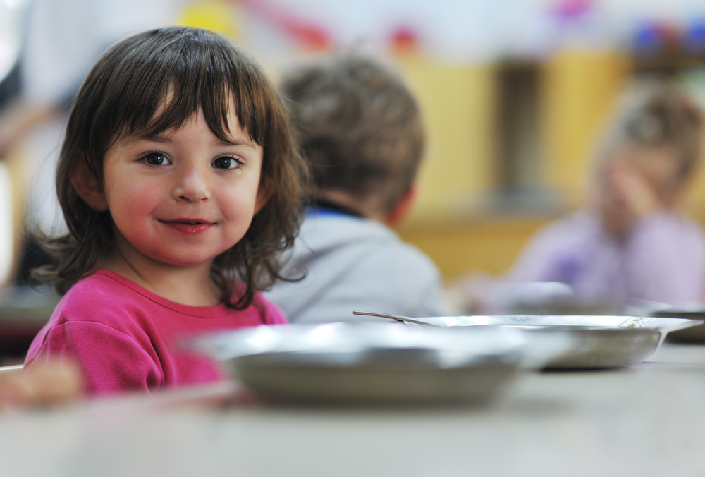 A young white girl with brunette hair wearing a pink shirt smiling at camera from the left side with a silver plate in front of her. The back of a blond boy's head is defocused in the background